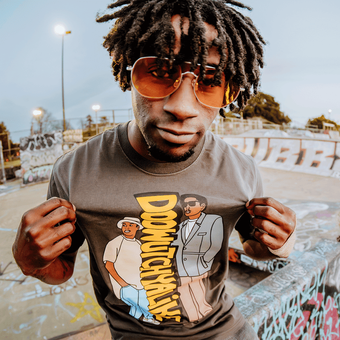 A person with short dreadlocks and amber sunglasses poses at a skatepark, holding out the Doowutchyalike Tee by Other, which features two illustrated people and the text “DOINGTHAN.” Graffiti covers the ramps and rails in the colorful background at dusk.