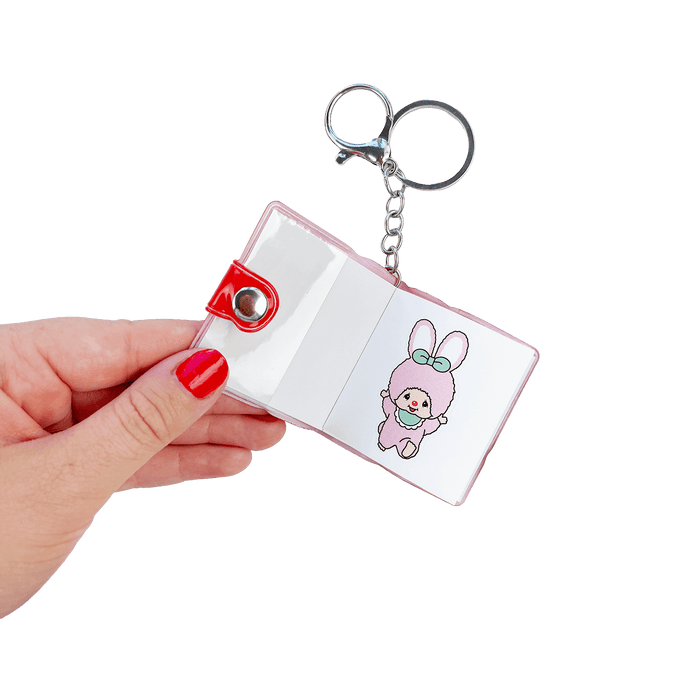 Hand holding open Monchhichi sticker book keychain featuring kawaii pink bunny character sticker — one of many cute designs inside this stationery girl bag charm