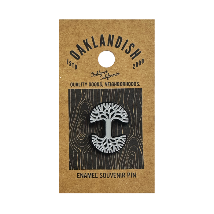 The Oaklandish Logo Enamel Pin by Oaklandish features a black and white enamel abstract tree on a brown kraft card reading "OAKLANDISH ESTD 2000, Oakland California, Quality Goods, Neighborhoods" with a grey woodgrain background.