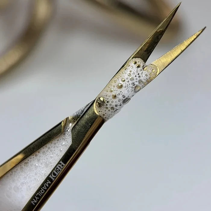 Close-up of gold-toned metal tweezers with a white foamy insert, showing the brand name 'RÉDI Marilyn' engraved on one arm against a soft light gray background.