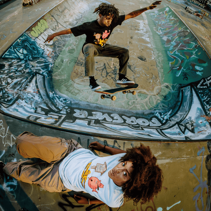 A woman with curly hair lies on graffiti-covered concrete, gazing up at the camera. Behind her, a man launches into a skateboard trick above a painted bowl. Both wear the Other brand’s Wet Since ’88 Heavy Tee—stylish hip-hop shirts that nod to Oakland heritage with bold cartoon faces. The colorful street art and lively urban energy make the scene dynamic and vibrant.