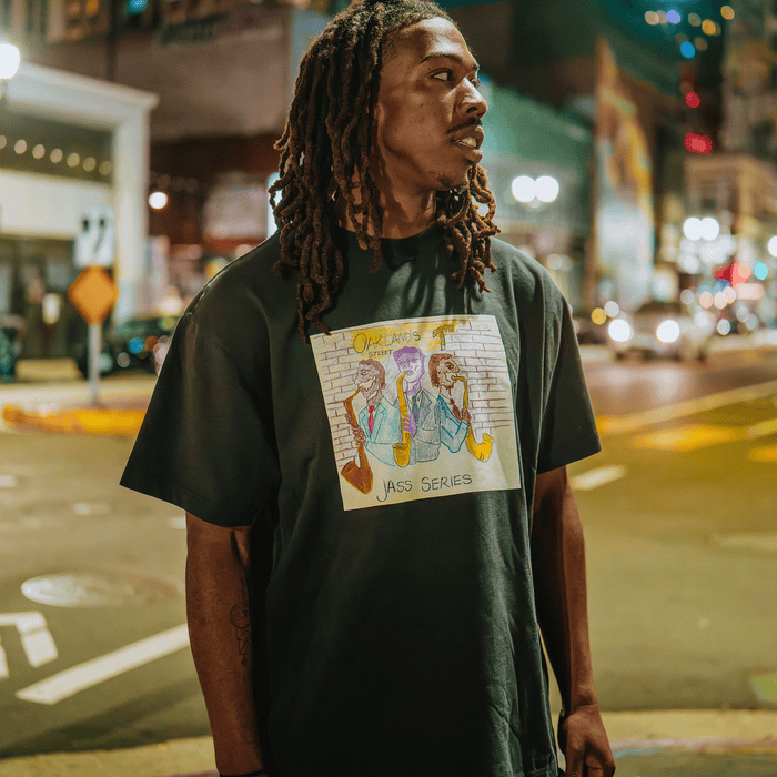 A man with long dreadlocks stands on 7th Street in Oakland at night, wearing the Other 7th St. Players Tee—a black shirt featuring a colorful cartoon graphic of three musicians. Blurred streetlights, buildings, and cars fill the background, capturing the lively spirit of Oakland jazz in this vibrant urban scene.