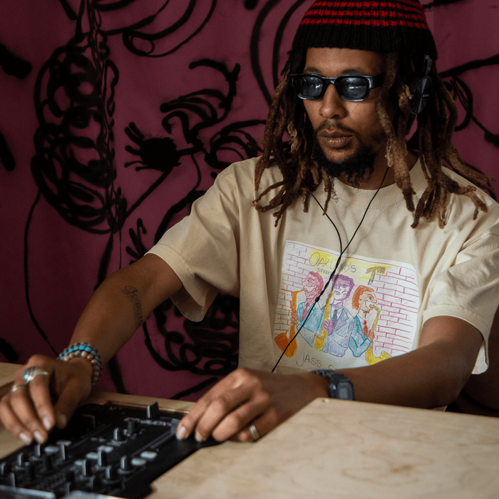 Wearing the Other 7th St. Players Tee, a person with dreadlocks, sunglasses, and headphones works the DJ controller. Their beige T-shirt features a vibrant cartoon graphic, paired with a red and black knit hat and beaded bracelets—an effortless nod to the creative energy of Oakland’s jazz scene. Behind them, a pink wall decorated with bold black abstract art evokes legendary venues like Chonto’s 7th St. Jass along historic 7th Street.