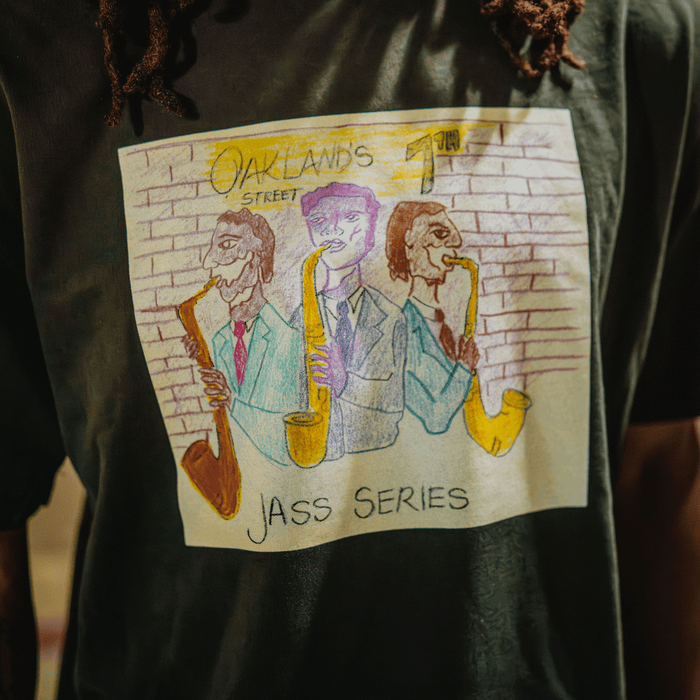 A person wears the Other 7th St. Players Tee, which features a vibrant illustration of three suited men playing saxophones against a brick wall. The text “Oakland’s Street Jass Series” celebrates the spirit of Oakland jazz on 7th street. The dark shirt draws attention to the colorful artwork, with only the torso visible in the image.