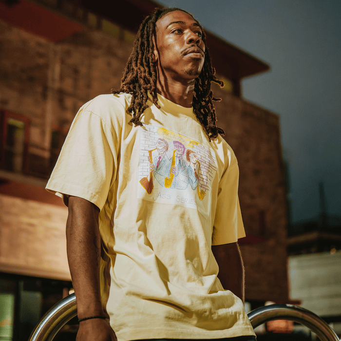 A man with long dreadlocks wears the 7th St. Players Tee by Other—a yellow t-shirt featuring a colorful cartoon graphic. He stands outdoors at dusk, gazing upward in front of a warmly lit building, capturing the vibrant spirit of Chonto’s 7th St. Jass and the iconic Oakland jazz scene. The background is softly blurred, highlighting him as the focal point.
