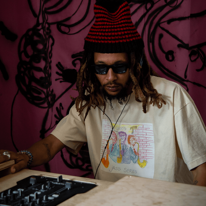 A man with dreadlocks, wearing sunglasses and a red-black beanie, sits at a DJ booth. He sports the 7th St. Players Tee from Other—a beige T-shirt adorned with a vibrant cartoon graphic. The pink backdrop, accented by bold black abstract tentacle-like designs, channels the lively spirit of Oakland jazz culture, calling to mind unforgettable nights on 7th street at iconic venues such as Chonto’s.