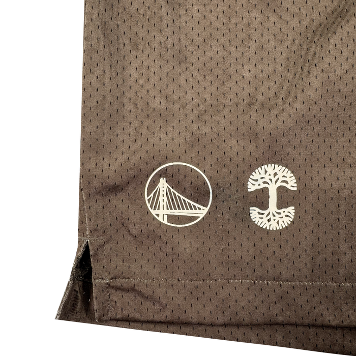 Close-up of a brown mesh fabric from the Arched Black-Letter Shorts by Other, featuring two white symbols: a stylized bridge inside a circle and a tree with roots and branches inside a broken circle. The Warriors x Oaklandish-inspired design includes a small side slit at the hem.