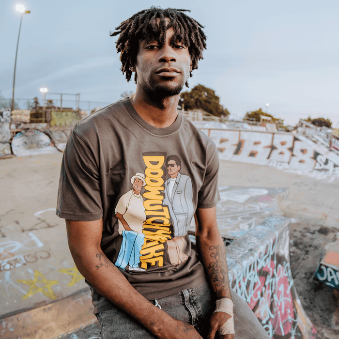 A man with short dreadlocks sits on the edge of a graffiti-covered skatepark ramp, wearing the Other Doowutchyalike Tee, which features cartoon characters and bold 90s hip hop-inspired graphics. He looks calmly at the camera; trees and fencing are visible in the background.