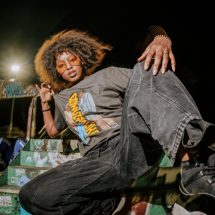 Wearing the Doowutchyalike Tee by Other, a person with curly hair, orange-tinted sunglasses, and baggy black jeans poses confidently on graffiti-covered stairs at night—one hand on their knee and the other resting behind them—as they look directly at the camera.