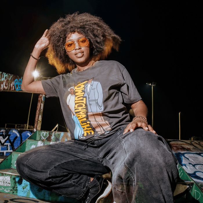 A person with a natural afro hairstyle and orange-tinted glasses smiles while sitting on graffiti-covered steps at night. They wear the Other Doowutchyalike Tee, dark jeans, and sneakers, with streetlights and a dark sky in the background.
