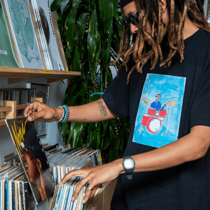 A person with dreadlocks and sunglasses browses vinyl records in a store, pulling out an album with a woman’s face on the cover. They wear Other’s Drummin’ Blues Tee, which features a painted drummer—a nod to Oakland jazz art—along with a watch and beads, surrounded by green plants and shelves of records.