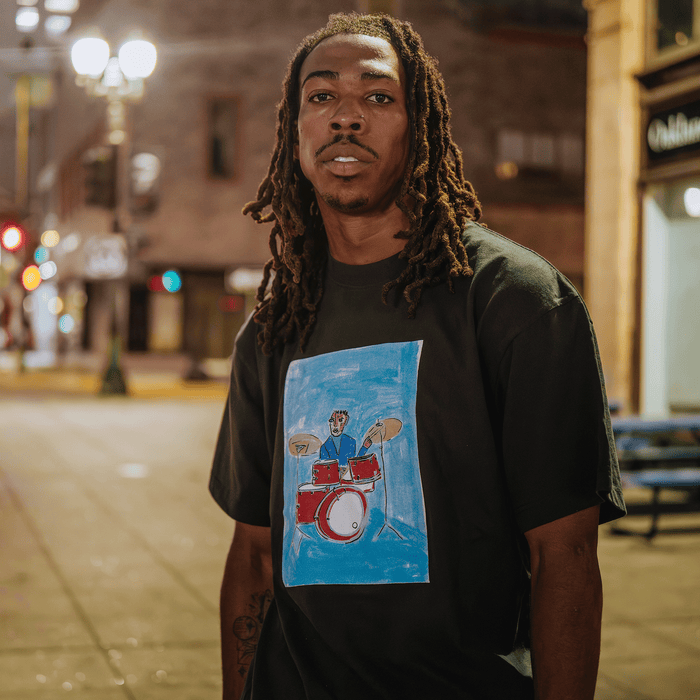 A man with long dreadlocks stands outside at night, wearing the Drummin’ Blues Tee by Other, which features a colorful illustration of a person playing drums. The background is filled with blurred city lights, benches, and buildings, evoking an urban nighttime vibe inspired by Oakland jazz art and West Oakland history.