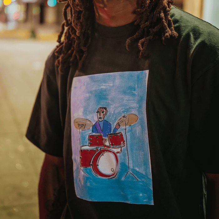 A person with long dreadlocks wears the Drummin’ Blues Tee by Other, which features a colorful illustration of a drummer on a red drum set against a blue background, capturing the vibrant spirit of Oakland jazz art. The photo is taken outdoors at night, softly lit by streetlights.