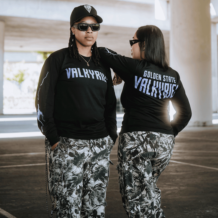 Two people pose confidently in the city, both wearing black GSV Golden State Valkyries Violet Lightning LS Tees and camouflage pants. The back of one shirt features "Golden State Valkyries" as they face away from the camera.