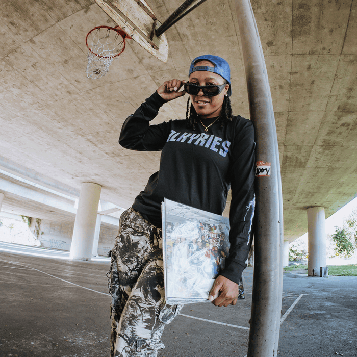 A person in a blue cap, sunglasses, and the GSV Golden State Valkyries Violet Lightning LS Tee made from 100% cotton leans on a pole under a highway overpass, holding a vinyl record. A basketball hoop and Valkyries graffiti color the pillars behind.