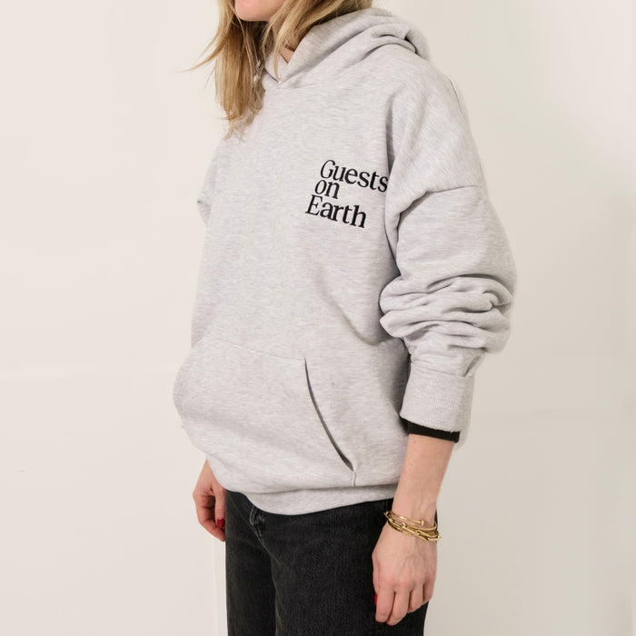 Woman wearing Guests on Earth Hoodie in heather grey, relaxed fit with bold chest logo and front pocket