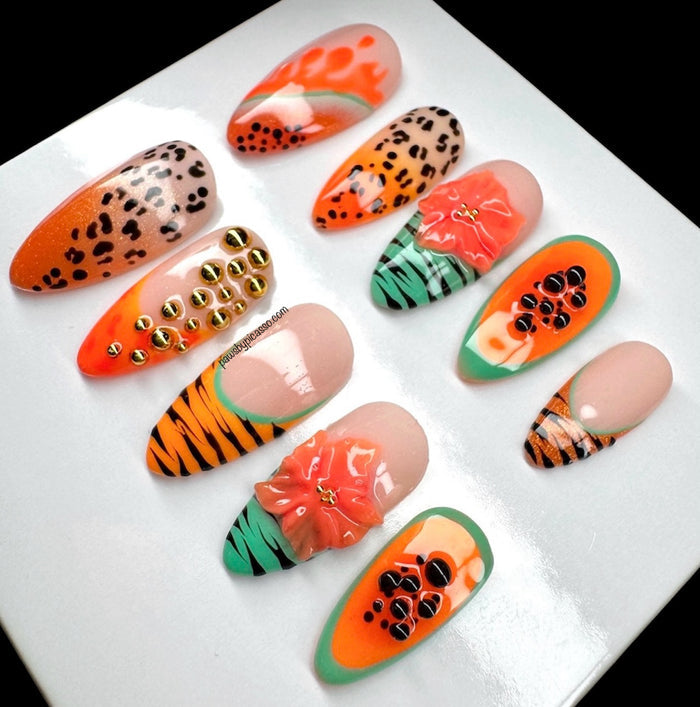 Ten decorative press-on nails displayed on a glossy white rectangular tray against a black background. The nails feature bold tropical and animal-inspired designs including orange and black leopard print, green and orange stripes, pink and orange floral patterns, and black seed details on an orange background.