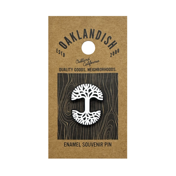 The Oaklandish Logo Enamel Pin by Oaklandish is a white enamel lapel pin shaped like an oak tree, attached to brown kraft paper backing and featuring text: “OAKLANDISH ESTD 2000,” “Oakland California,” and “ENAMEL SOUVENIR PIN.”.