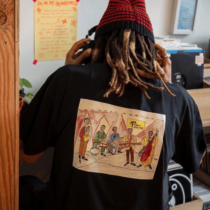 Wearing the classic fit Ode to Jass Tee by Other, which features a colorful band illustration on the back, a person with long, light brown dreadlocks adjusts their headphones in a cozy room filled with records, handwritten notes, a wooden shelf, and a red-striped knit hat—a space that reflects their deep connection to the vibrant Oakland jazz and early jazz scene.