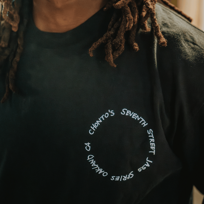 Close-up of a person wearing the Ode to Jass Tee by Other—a classic fit black t-shirt with white curved text reading “CHONTO’S SEVENTH STREET SERIES OAKLAND, CA.” The image shows the person’s neck, part of their shoulder, and brown dreadlocks against a softly blurred background that echoes the spirit of Oakland’s early jazz scene.