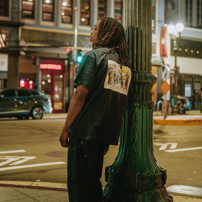 At night, a person with long dreadlocks leans against a green streetlamp, wearing the Other brand’s Ode to Jass Tee—a classic fit t-shirt featuring a vibrant illustration on the back. City lights, cars, and buildings line the lively Oakland street in the background, capturing the energetic spirit of jazz.
