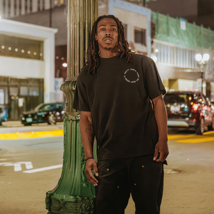 A man with long dreadlocks and a beard leans against a green lamppost at night on a city street, channeling the laid-back spirit of the Oakland jazz scene. He wears Other’s Ode to Jass Tee in a loose, classic fit, paired with black pants. Cars and illuminated storefronts glow in the background under streetlights.