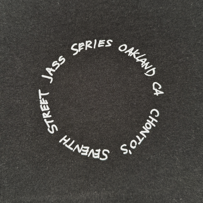 White text curves in a loose circle on black fabric, reading: “OAKLAND CA, CHONTO’S, SEVENTH STREET JASS SERIES.” The unevenly spaced letters evoke a hand-drawn aesthetic and capture the energetic essence of Oakland’s early jazz culture. The weave of the classic-fit Ode to Jass Tee by Other is visible, making this t-shirt a fitting tribute to the city’s enduring jazz legacy.
