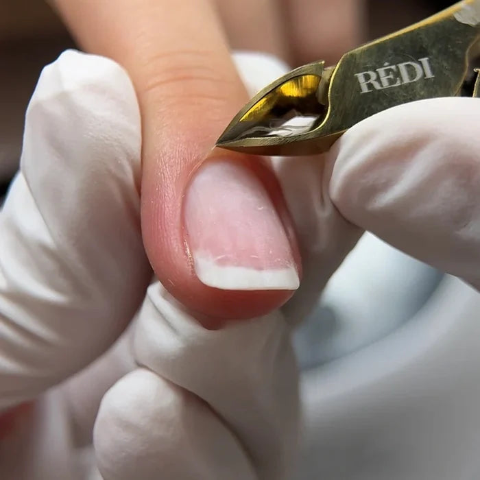 A close-up of a cuticle  being clipped with a gold REDI brand manicure tool, held by a hand wearing a white glove.