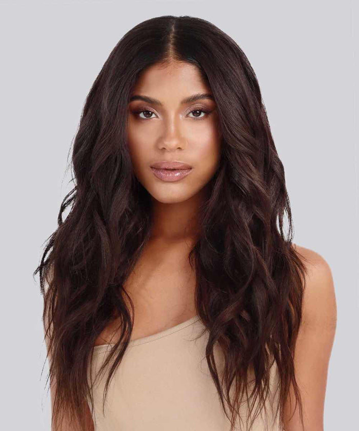 A woman with long, wavy black hair in Relaxed Straight extensions and glowing makeup looks ahead, wearing a beige off-shoulder top.