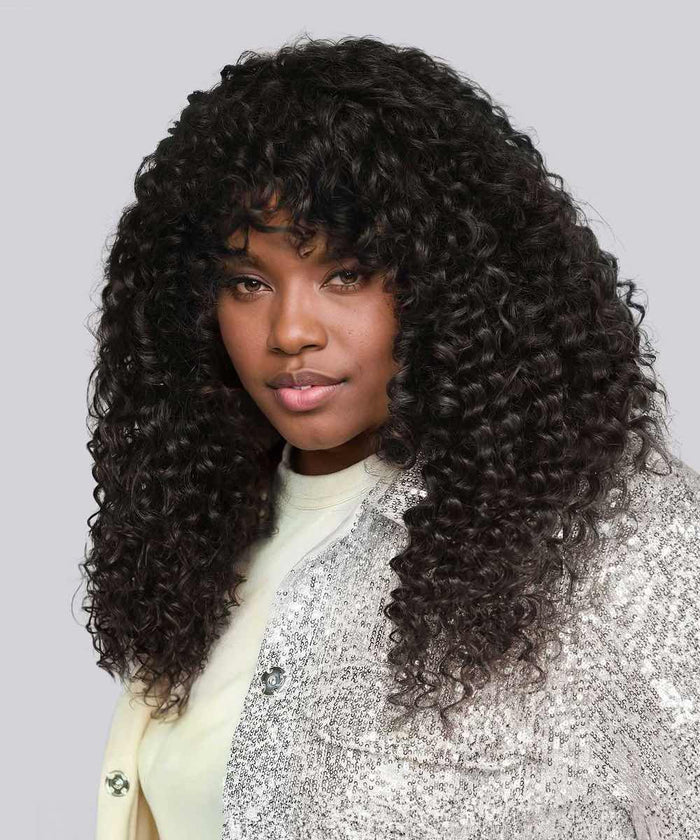 A woman with long, voluminous tight curls in Natural Black  wears a light top and sparkling silver jacket on gray background.