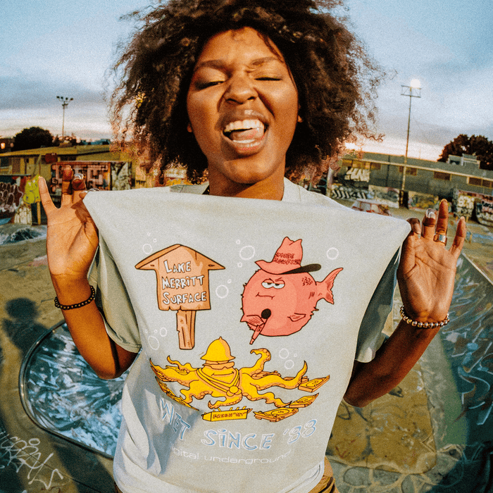 A joyful person at an Oakland skate park closes their eyes and sticks out their tongue while lifting their shirt to reveal the Other Wet Since ’88 Heavy Tee, which features a colorful hip-hop graphic of a fish, a “Lake Merritt Surface” sign, and yellow creatures. Graffiti and ramps fill the background.