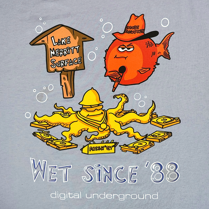 On the Wet Since ’88 Heavy Tee by Other, a cartoon orange fish in a cowboy hat smokes a pipe by a “Lake Merritt Surface” sign, capturing true Oakland flair. A funky yellow octopus armed with vinyl records wraps around an iconic Oakland fire hydrant. Playful hip-hop-inspired text underneath declares: “Wet since ’88, digital underground.”.